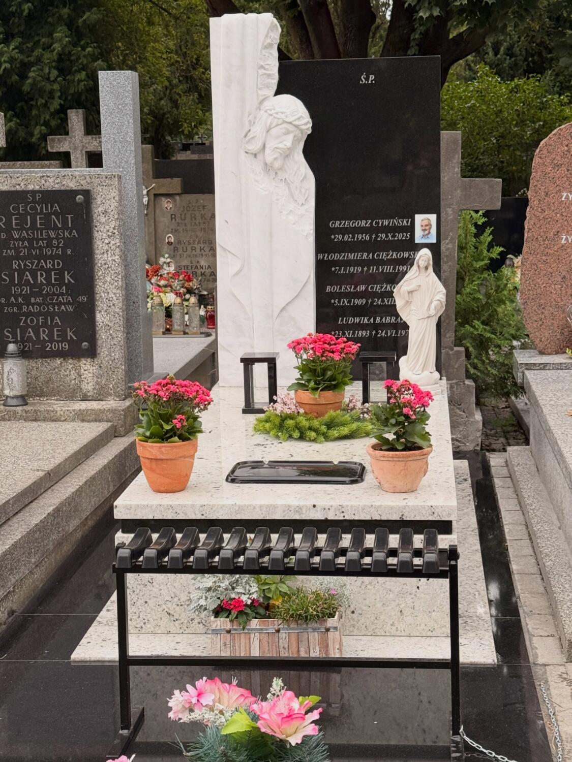 Trzy doniczki różowych kwiatów i dwie ciemne latarnie na jasnej płycie nagrobka