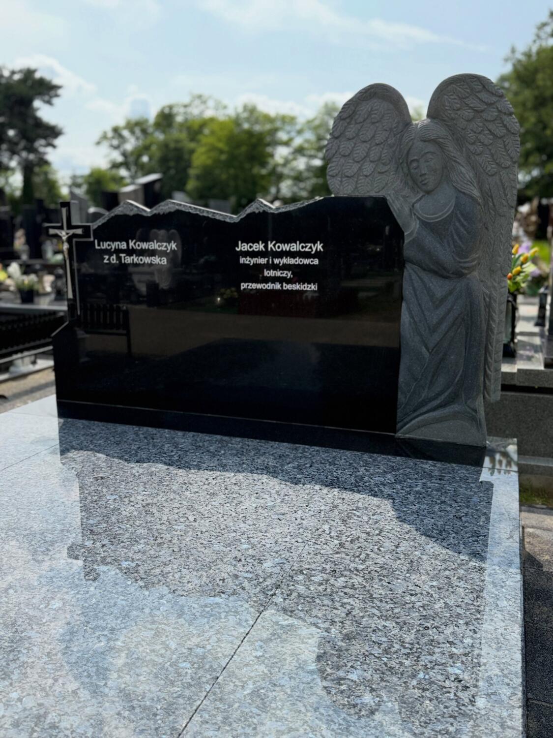 Dwa pola nazwisk na szerokiej ciemnej tablicy, krzyż po lewej i postać anioła po prawej — GRAAL